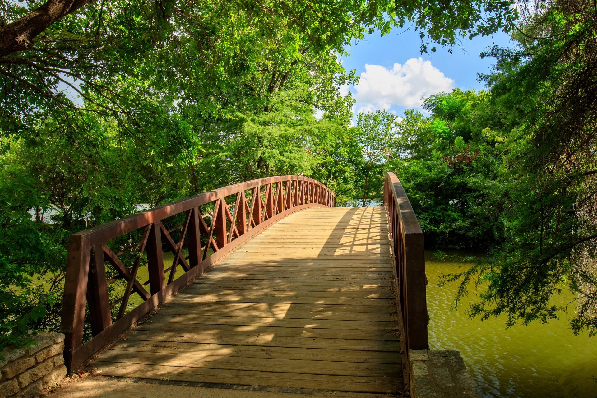 A wooden footbridge spans a city park trail in Austin, Texas, surrounded by lush green trees.