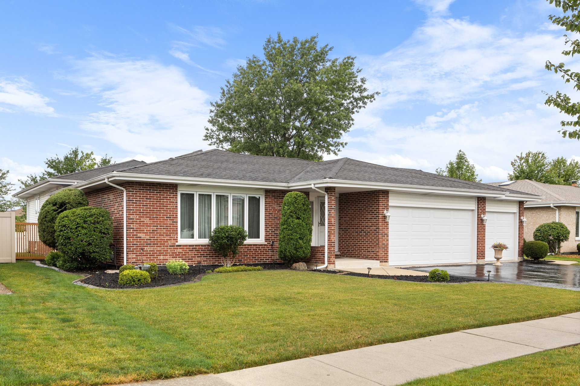 A suburban home with brick siding, a three car garage, neat landscaping, and a wet blacktop driveway