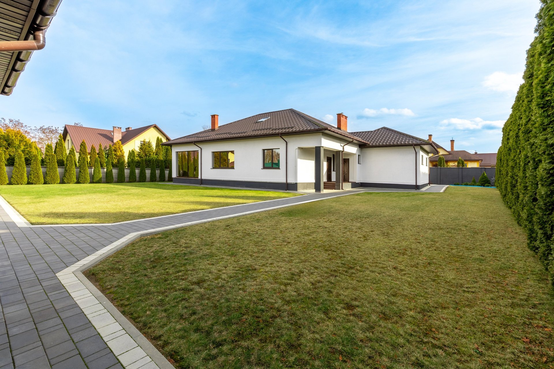 A modern suburban house with a brown roof, large green lawn, and a paved walkway, surrounded by a well-maintained hedge.