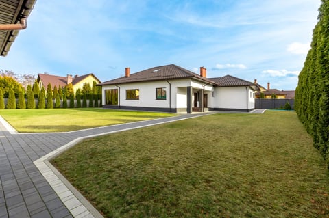 A modern suburban house with a brown roof, large green lawn, and a paved walkway, surrounded by a well-maintained hedge.