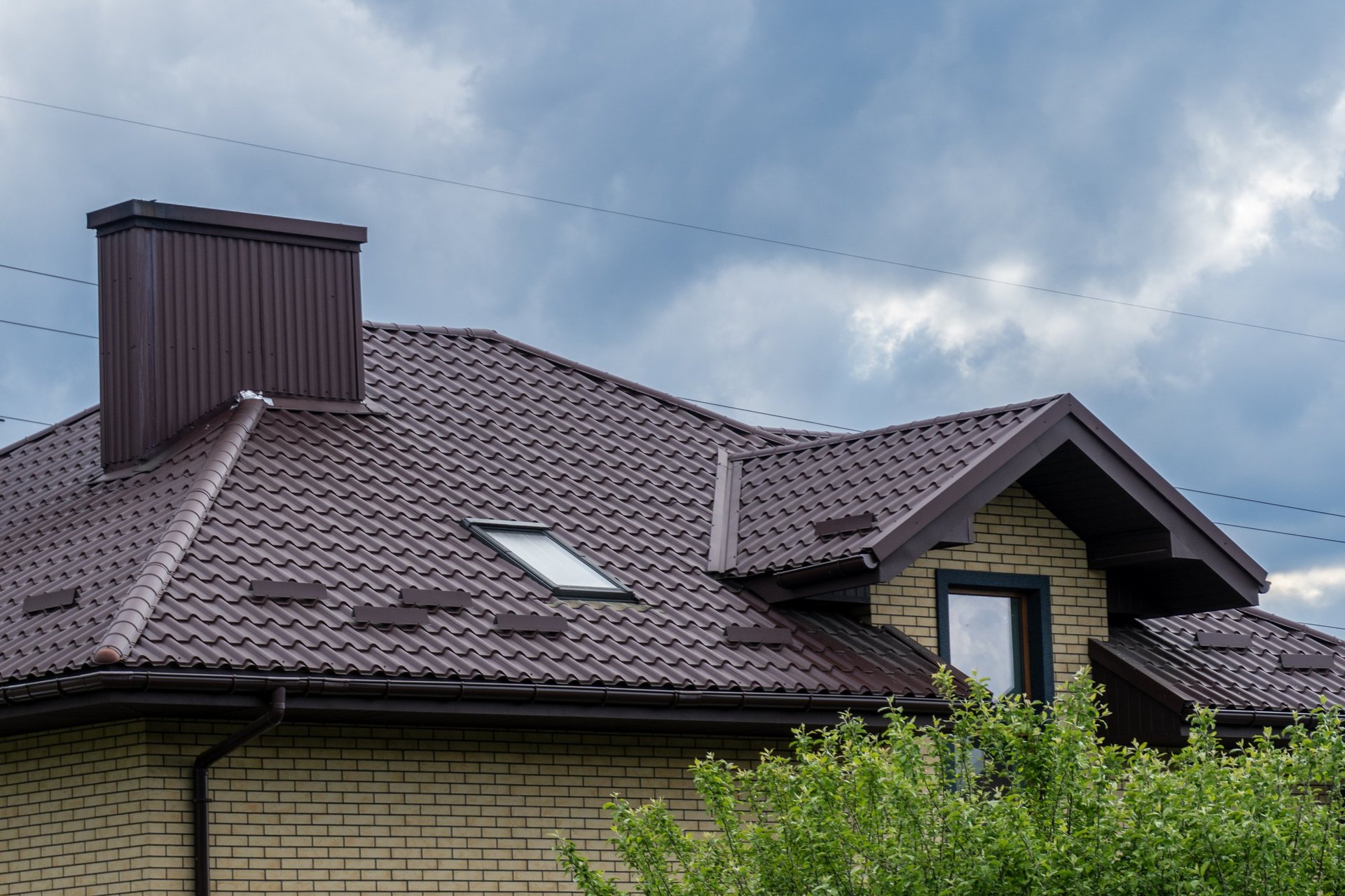 View of the top of a residential brick home featuring brown metal roofing, chimney, roof window, gutters, and cloudy sky