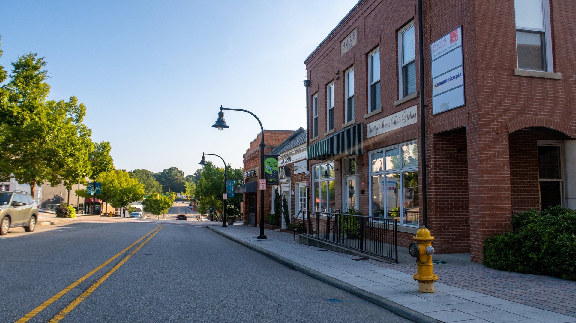 A quiet morning view of historic downtown with classic brick storefronts, tree-lined sidewalks, and vintage street lamps under clear blue skies