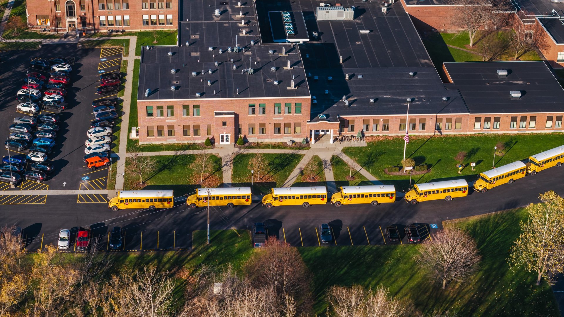 Aerial shot of an American school campus with yellow school buses lined up outside, surrounded by autumn trees and suburban greenery