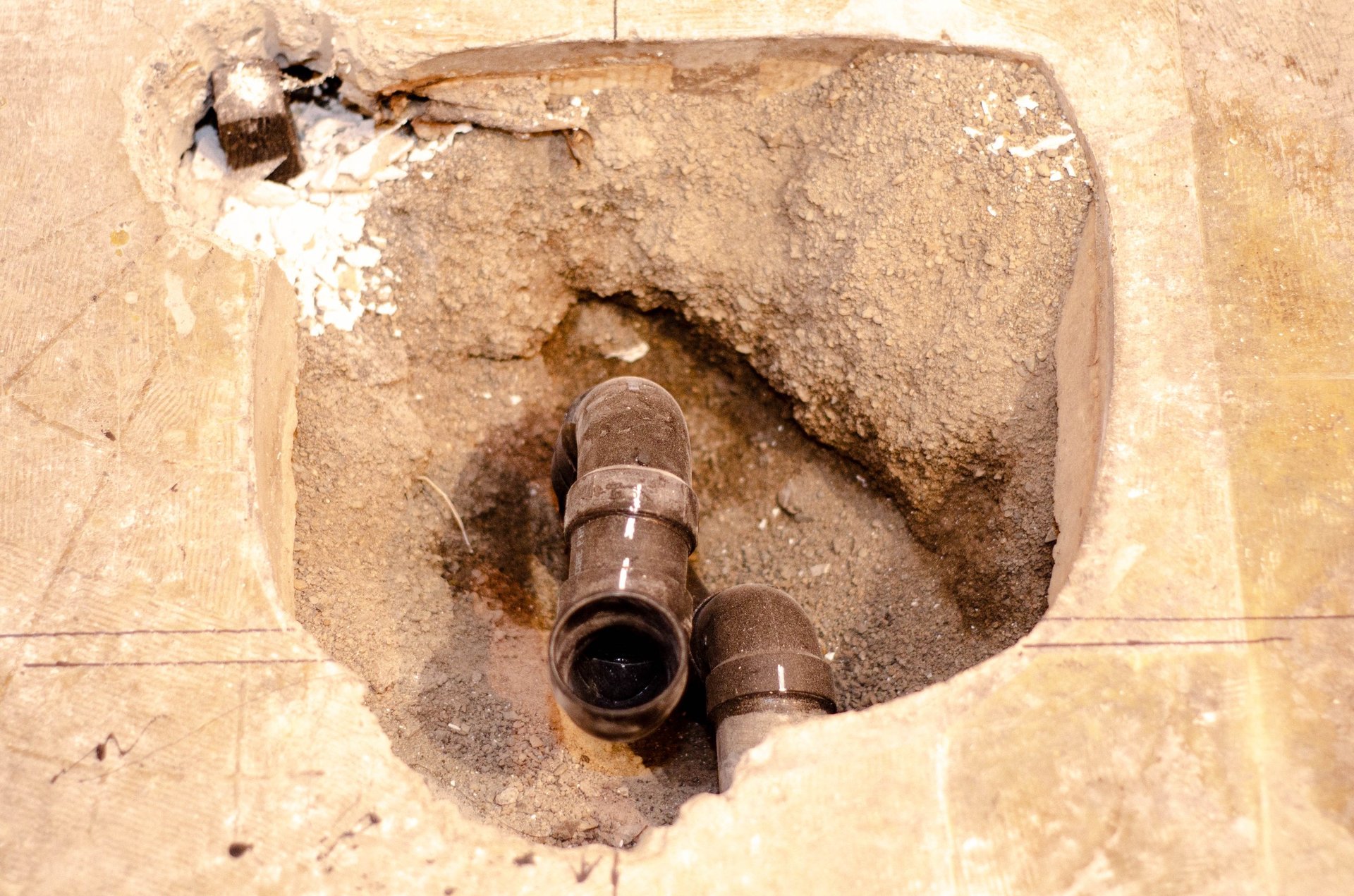 Open concrete floor revealing plumbing pipes during renovation work. The image shows drainage lines, construction debris, and infrastructure access for repair, installation, or maintenance in a building foundation