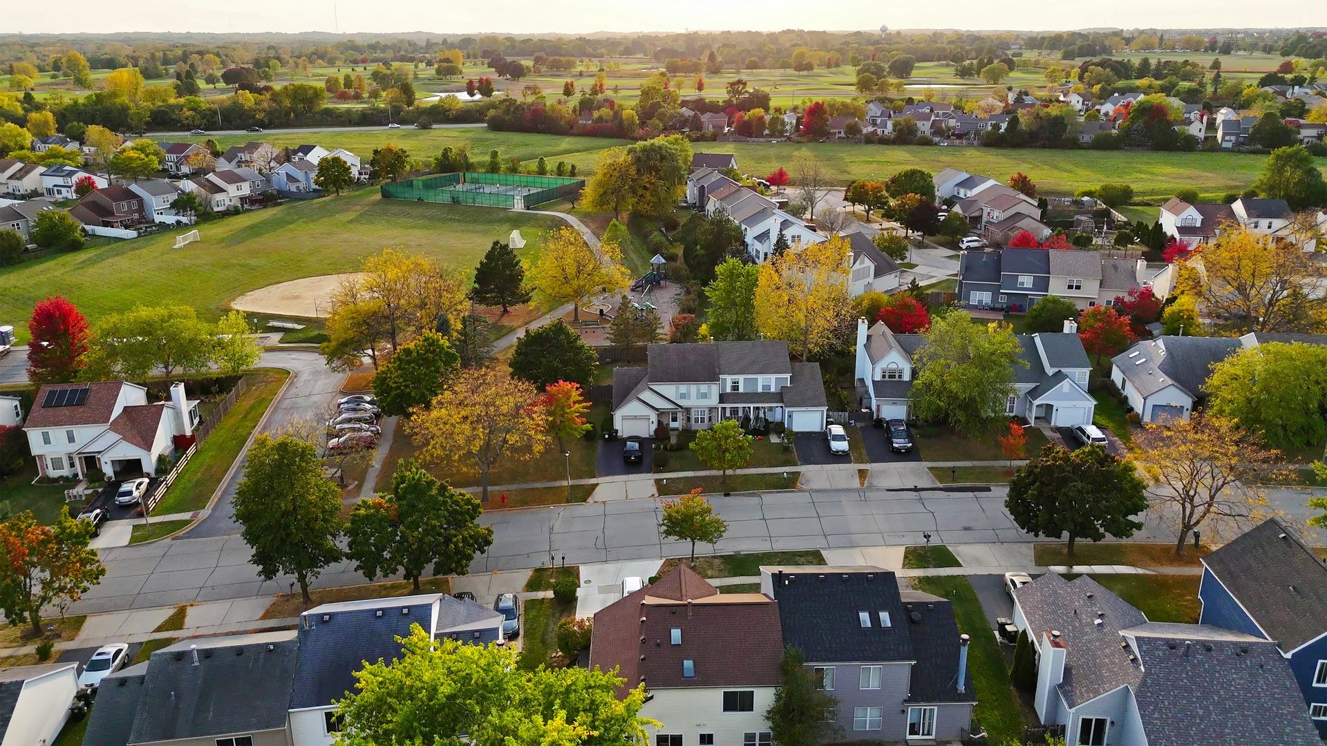 A high-angle aerial view of a suburban neighborhood in Chicago, Illinois, featuring residential homes, tree-lined streets, parks, and open green spaces.