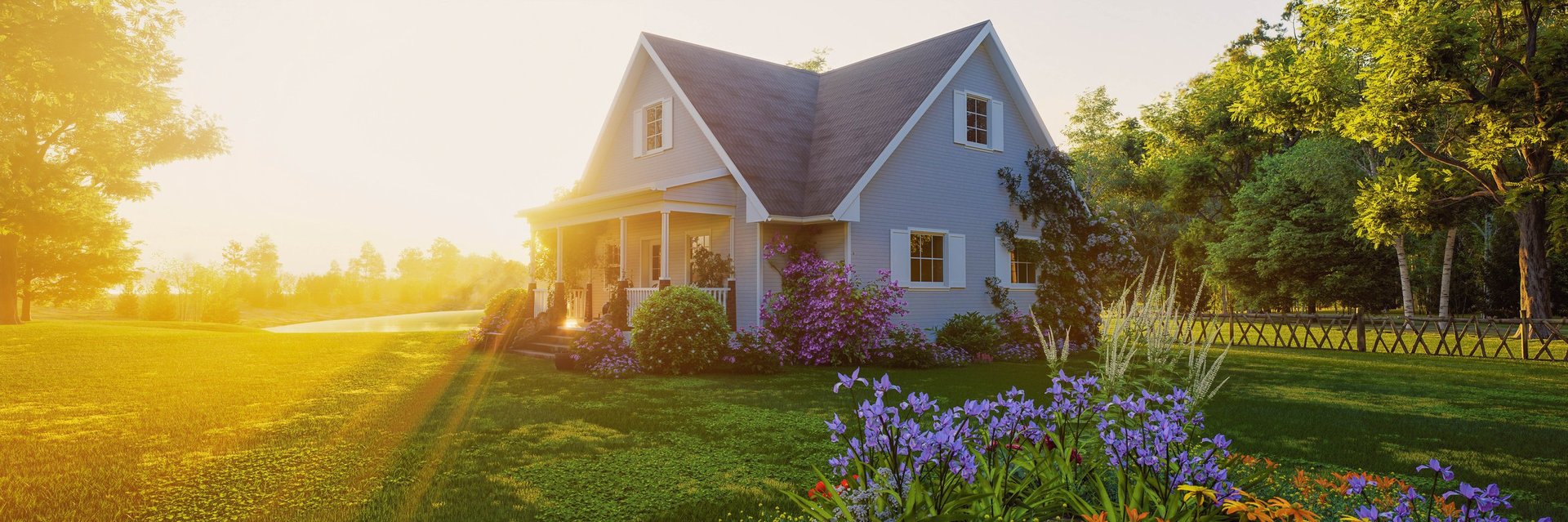 A small house with a porch sits in a lush green yard surrounded by blooming flowers, trees, and a wooden fence under warm sunset light