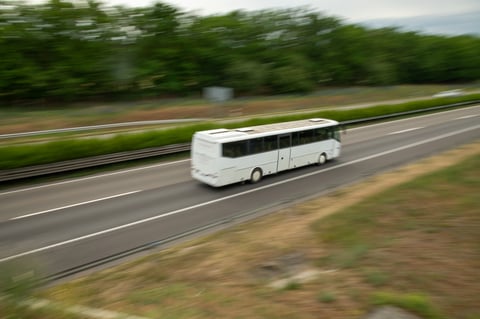 A white bus moves quickly on a highway. The road stretches ahead while green trees line the sides. Clouds fill the sky as the bus continues its journey.