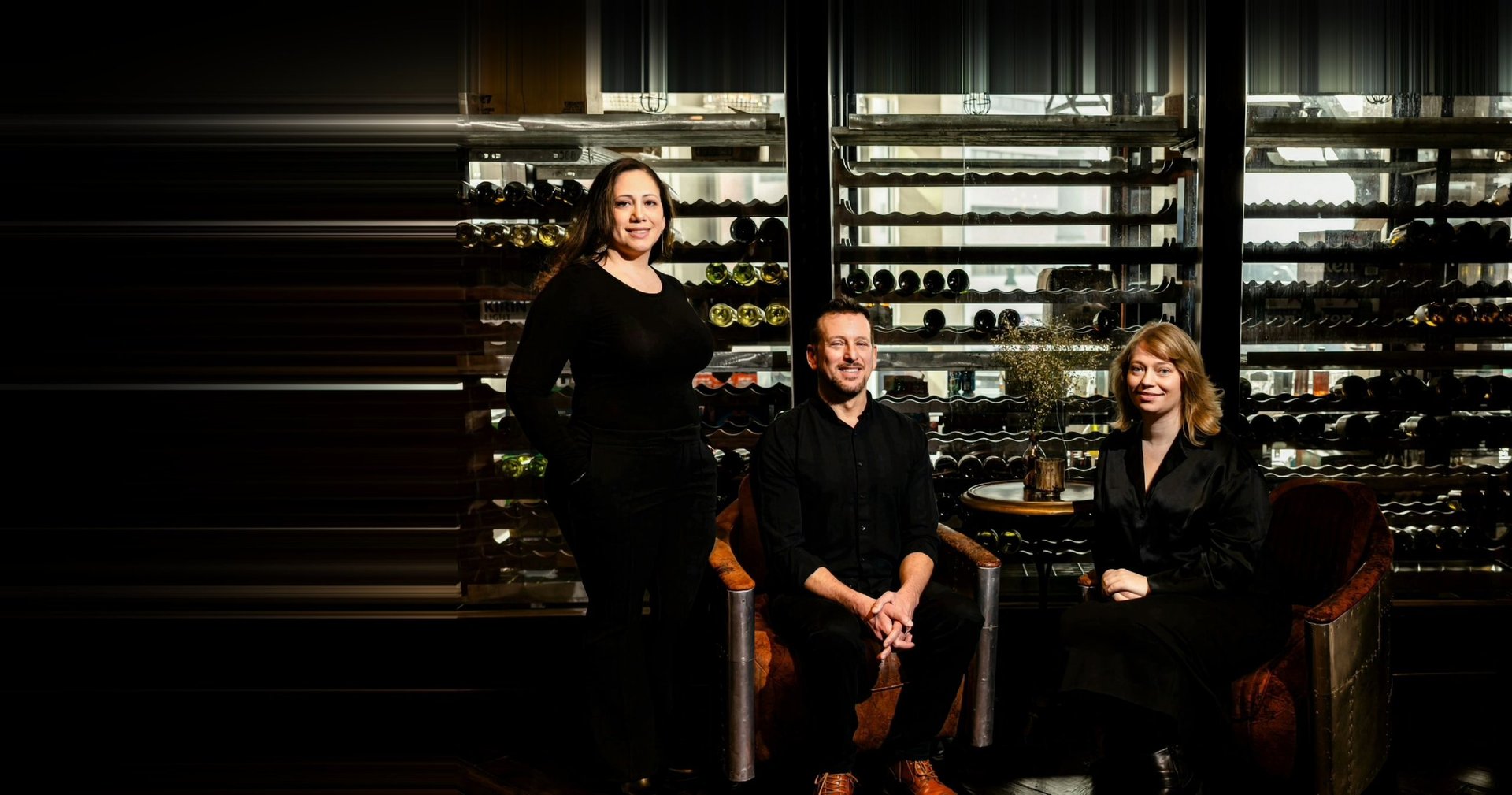 Three musicians in dark suits posing with string instruments against an illuminated wine rack backdrop