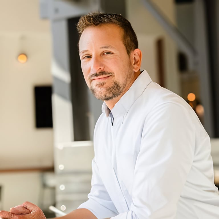 Man with brown hair and goatee wearing light blue button-up shirt, smiling at camera in modern interior setting