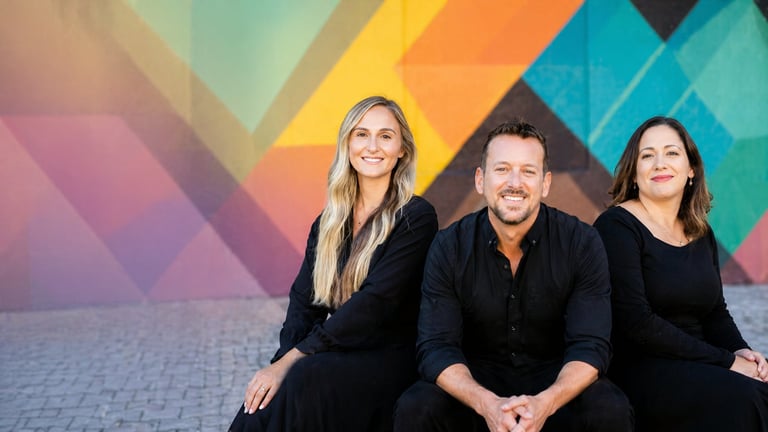 Three professionals in dark clothing sitting together against a colorful geometric background with diagonal shapes