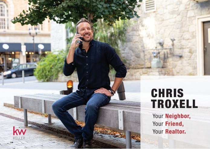 Chris Troxell, a real estate agent, sitting on a bench in a courtyard wearing a dark shirt and jeans, with Keller Williams branding visible